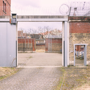 Gesicherter Gefängniseingang mit Metalltor und Stacheldraht, der in einen leeren Innenhof zwischen Backsteinmauern führt.