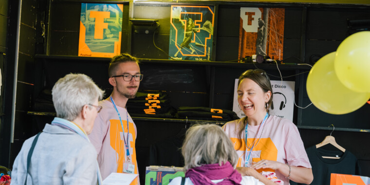 Infobox des Festival Theaterformen. Vier Menschen, zwei in der Box, zwei vor der Box agieren miteinander. Die eine Person lacht. Im Hintergrund sind Poster zu sehen, gefaltete T-Shirts und an der Seite stehen Programmhefte und Ballons hängen rechts in der Ecke.