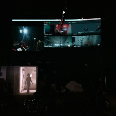 A dark room; above, several live video images are shown on a large screen. Below, a door opens through which a person, visible only as a silhouette, steps into a lit room. The scene conveys a controlled, observant atmosphere.