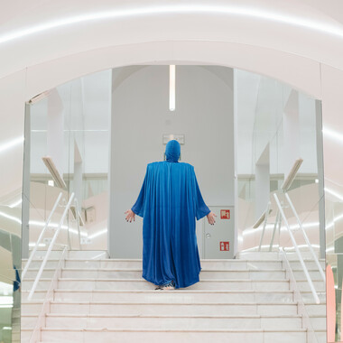 A white staircase bordered on the left and right by mirrored walls. A person stands in the middle of it. The person can be seen from behind and is dressed from head to toe in a long-sleeved, flowing robe in the color blue. Both arms are slightly outstretched, the open palms point towards the direction of the white room to which the staircase leads.