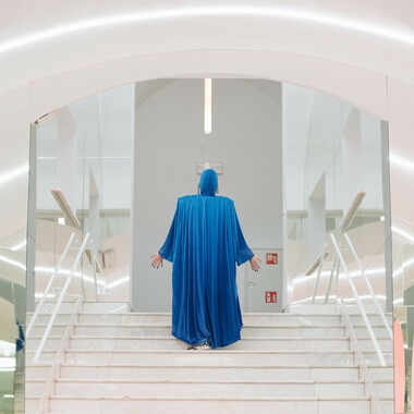 Eine weiße Treppe, die links und rechts von verspiegelten Wänden eingegrenzt wird. Auf dessen Mitte steht eine Person. Sie ist von hinten zu sehen und steckt von Kopf bis Fuß in einem langärmeligen, fließenden Gewand in der Farbe Blau. Beide Arme sind leicht ausgestreckt, die offenen Handinnenflächen zeigen in die Richtung des weißen Raums, zu dem die Treppe führt.