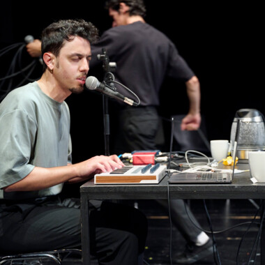 A person is sitting on stage at a table with a laptop, MIDI keyboard, and microphone, singing with their eyes closed. In the background, two other people are moving, partially out of focus. On the table are white mugs and a thermos.