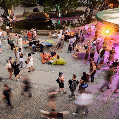 A bird's eye view of the Festival Theaterformen festival center. Many people in one area, some standing and sitting, others walking past. Motion blur can be seen in the right half of the picture.