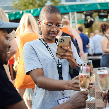 Eine Person mit Brille im Festivalzentrum unter freiem Himmel, die ein graues T-Shirt und ein schwarzes Halsband mit dem Festival Theaterformen-Logo trägt. Die Person fotografiert mit dem Handy das volle Sektglas, dass die Person in der rechten Hand hält. Im Hintergrund sind andere Festivalbesucher*innen von hinten zu sehen.