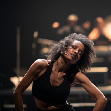 Ein Schwarzweiß-Foto aus dem Stück “When I saw the Sea” von Ali Chahrour: Ein*e Performer*in mit dunklen, langen, welligen Haaren an einem Marmortisch, der*die in die Kamera blickt. Auf der Marmorplatte vor ihr*ihm ist die Reflektion der Person zu sehen.
