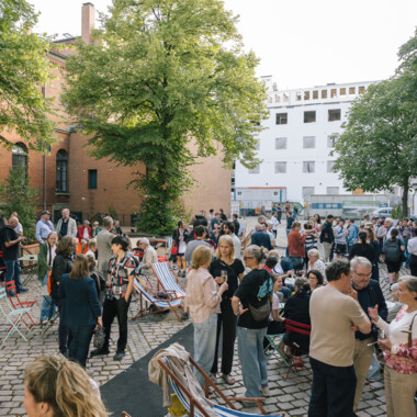 A large group of people gather outside on a paved square between trees and buildings. The atmosphere is lively and sociable - many people are standing in groups, chatting or drinking. A red brick building and a modern white building can be seen in the background. A few colorful folding chairs and deckchairs are scattered around the square, giving the place a relaxed ambience.