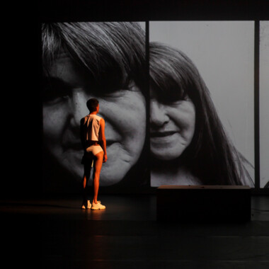 Bühnensituation: Tiziano Cruz steht mit dem Rücken zum Pubikum und schaut auf die Projektion eines Gesichts.