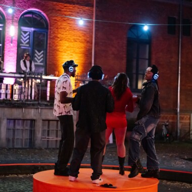Four people dance at an outdoor silent disco at night, standing on an orange platform. They are wearing glowing headphones. In the background, two DJs stand at a booth on a balcony in front of a colorfully lit brick facade.