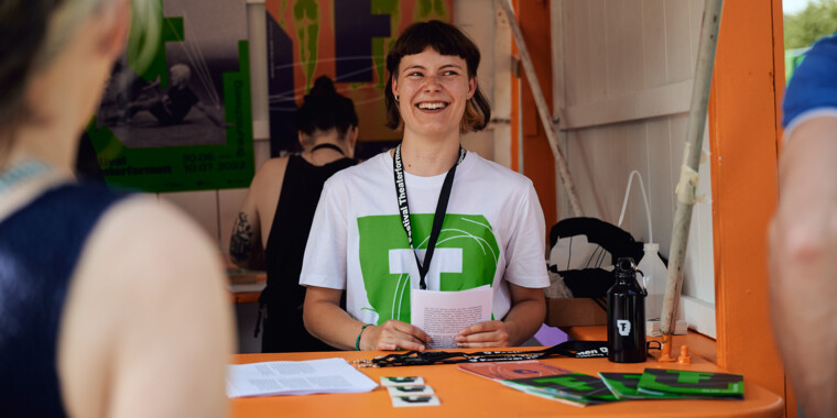A person in a white T-shirt with a green Theaterformen logo standing in the info box and smiling. The person has chin-length dark hair with bangs and is holding a pile of notes in their hands.