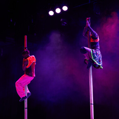 Zwei Artist:innen in bunten Kostümen klettern an langen vertikalen Stangen nach oben. Die Szene ist in buntes Licht und Nebel getaucht.