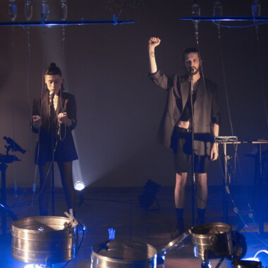 Zwei Personen stehen auf einer Bühne vor Mikrofonen, umgeben von technischen Geräten und von der Decke hängenden Infusionsflaschen. Eine Person hebt die Faust. Die Szene ist schwach, bläulich beleuchtet.