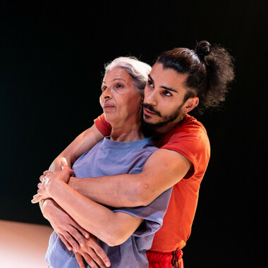A dancing couple with a big age gap. The older person is Mimouna Latifa Khamessi. Behind her stands her son Mohamed Toukabri, who embraces her gently. She is wearing a blue tshirt, her gray hair is tied back. Mohamed wears a red tshirt and has long, dark curly hair tied up into a high ponytail. Both are looking into the distance.