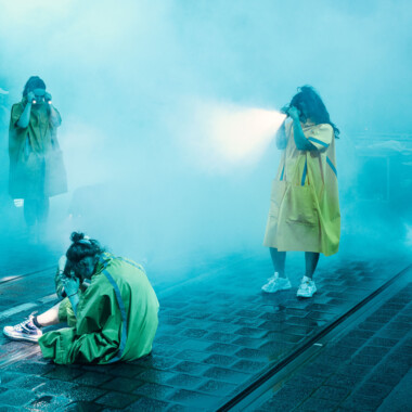 Eine Performance im öffentlichen Raum. Drei Personen in gelben Regenponchos befinden sich auf einer gepflasterten Straße umgeben von viel Nebel. Zwei stehen, sie halten Taschenlampen an die Schläfen, sodass ihre Blickrichtung beleuchtet wird. Eine Person sitzt und betrachtet den Boden. Die Szene ist bläulich beleuchtet.
