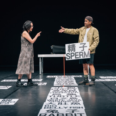 Zwei Personen stehen sich auf einer Bühne gegenüber und gestikulieren, während einer ein Schild mit der Aufschrift SPERM und chinesischen Zeichen hält. Auf dem Boden liegen zahlreiche Karten mit englischen und chinesischen Begriffen verteilt.