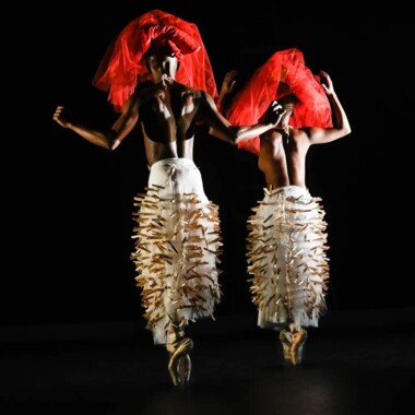 Ein Foto von dem Stück "Hatched Ensemble": Die Rückenansicht von zwei Tänzer*innen auf der Bühne. Beide sind oberkörperfrei und tragen weiße lange Röcke, die mit hölzernen Wäscheklammern versehen sind. Sie tanzen auf den Fußspitzen und tragen auf ihn Köpfen Schleier aus Tüll in der Farbe rot.