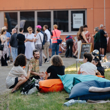 Besucher*innen unterhalten sich auf einer Wiese im Festivalzentrum. Einige sitzen auf bunten Sitzsäcken, andere stehen in kleinen Gruppen vor dem Eingang und kommen miteinander ins Gespräch.