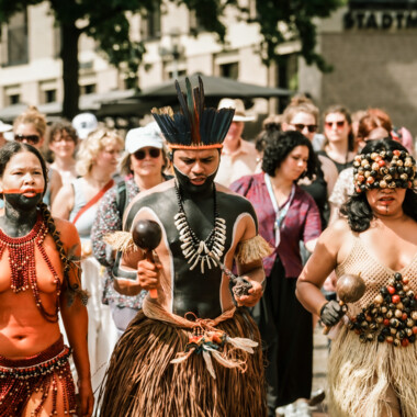 Three people in traditional attire lead a procession through a crowd.
