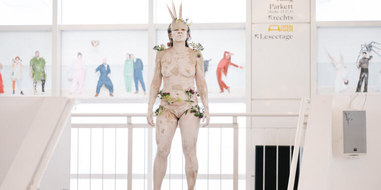 A person, painted and adorned with plants and feathers, stands at the top of a staircase in a bright foyer.