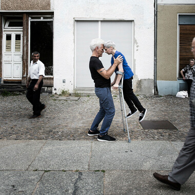 Self-description of the depicted persons: Two performers standing in the middle of a sidewalk, forehead to forehead. On the right is Claire Cunningham, a short, white woman in her early forties.  She has short white hair and is wearing a blue t-shirt and black trousers and shoes.  Claire is leaning on grey elbow crutches so that her feet are not touching the ground. On the left is Jess Curtis, a tall, white man in his mid-fifties with an athletic build.  He has short white spikey hair and is wearing grey trousers and a black t-shirt and shoes. Jess is holding the woman by her shoulders. Passersby can be seen in the background and foreground.
