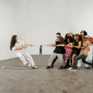 A group of people are pulling on a rope. On the left, a single woman, dressed entirely in white, is pulling hard on the rope, leaning backwards as she does so. On the right, a group is pulling in the opposite direction. The scene is set in a simple room with white walls and a tiled floor.