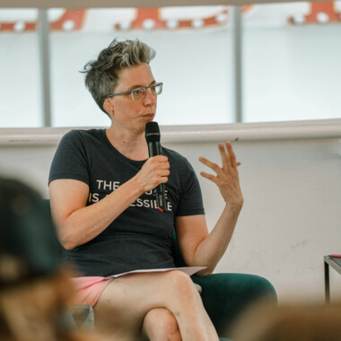 Eine Person sitzt in einem grünen Sessel, spricht in ein Mikrofon und gestikuliert mit der linken Hand. Sie trägt eine Brille, ein dunkelblaues T-Shirt mit der Aufschrift „THE FUTURE IS ACCESSIBLE“ und rosa Shorts. Im Vordergrund sind unscharf Köpfe des Publikums zu sehen.