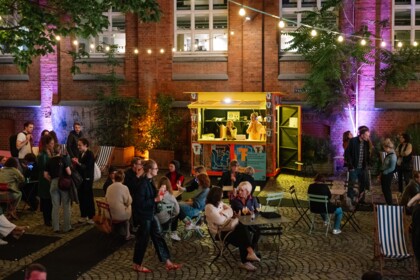 Das Festivalzentrum im Theaterhof bei Nacht. Menschen sitzen und stehen in kleinen Gruppen beisamen, unterhalten sich und trinken etwas. Im Hintergrund ist die Infobox des Festivals. Lichterketten hängen zwischen den Bäumen.