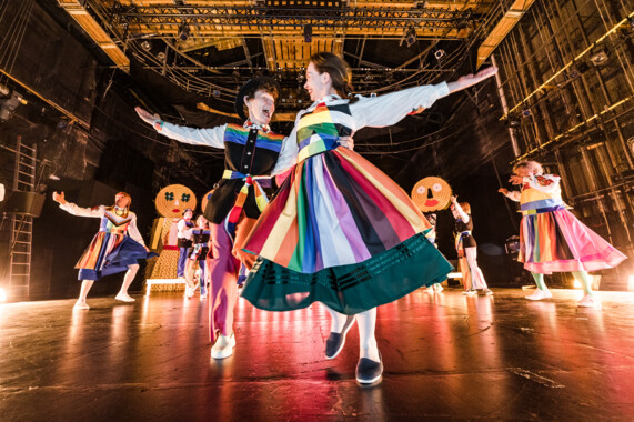 Stage situation: Many cheerful people in colourful traditional clothing dance together or alone.