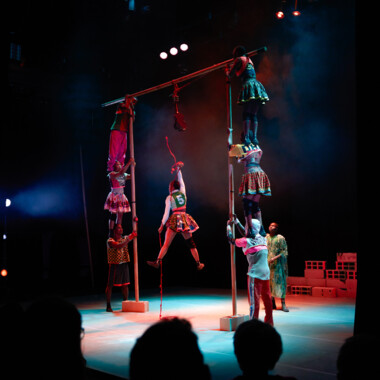 Mehrere Artist:innen auf der Bühne. Aus 3 langen Stangen eine Konstruktion gebaut, zwei vertikale Stangen sind oben durch die dritte verbunden. Jeweils 3 Personen stehen an jeder Seite übereinander, um die Konstruktion zu fixieren Eine Person hängt in der Mitte an einem Seil, das Publikum im Vordergrund im Dunkeln sitzt.