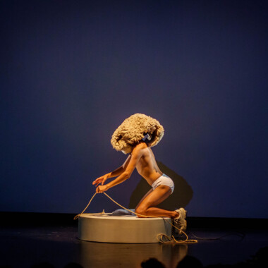 Stage situation: Tiziano Cruz kneels on a platform in his underwear and wearing a sheepskin. He holds a rope like reins.