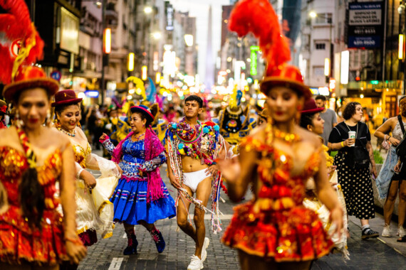 Several people in colourful, glittering costumes on a street full of pedestrians. In the middle, Tiziano Cruz in underwear and also covered in colourful fabrics.
