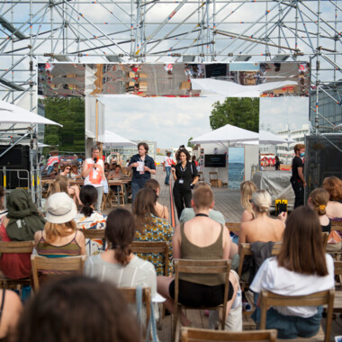 Studierende des Festivalcampus Niedersachsen während eines Gesprächsformat beim Festival Theaterformen. Sie sitzen draußen auf Holzstühlen, vorne stehen drei Personen, von denen eine in ein Mikrofon spricht. 