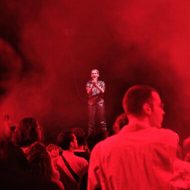 A concert situation. In the foreground, a standing audience can be seen from behind. Ivana Jozic is on stage, holding a microphone with both hands, Ivana's mouth is open. Ivana has pulled her short blonde hair into a deep side parting and is wearing a short-sleeved jumpsuit made of dark leather. The stage is covered in fog.