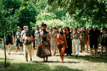 Eine Szene aus dem Spaziergang entlang des Leineufers "We Are Rivers". Eine Gruppe in der Natur umgeben von Bäumen und Wiese. Im Vordergrund sind drei Personen in auffälligen Kostümen, denen die Gruppe hinterher läuft.