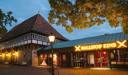 In the photo is the Ballhof Eins building with a sign saying Ballhof Eins. The yellow lettering is lit brightly. The sign is above the entrance, which is glazed. There is a lantern in front of the building which is also lit. 