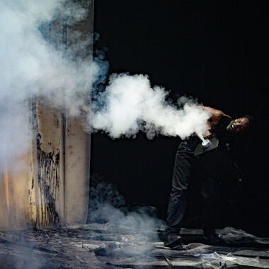 Bildbeschreibung seitens der Festival Theaterformen Redaktion. Selbstbeschreibung der abgebildeten Personen folgt. / Eine Person mit einer Nebelmaschine, aus der Nebel kommt, steht auf der rechten Seite. Sie ist nach vorne gebeugt und schaut nach links. Sie trägt eine schwarze lange Hose und ist oberkörperfrei. Der Hintergrund ist schwarz, nur auf der linken Seite sieht man drei weiße Stoffstreifen, von denen schwarze Farbe runtertropft.