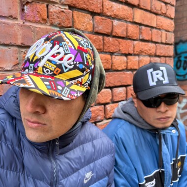The hip-hop duo Eskina Qom: They are sitting in front of a brick wall and wearing sporty clothes. The person on the left is looking to the left and is wearing a colorful cap and a dark blue Nike jacket zipped up high. The person next to them has dark sunglasses on their nose and is wearing a dark cap with the letter R on it. The person is wearing a blue jacket with a gray hoodie underneath.
