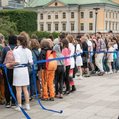 Eine Performance im öffentlichen Raum. Eine große Gruppe von Menschen steht dicht zusammen auf einem Platz hinter einem langem blauen Absperrband, dass sie scheinbar zusammenhält. 