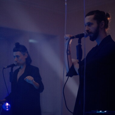 Zwei Personen stehen in einem dunklen, blau beleuchteten Raum auf einer Bühne. Sie tragen dunkle Kostüme und sprechen in Mikrofone. 