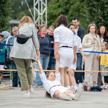 Eine Performance im öffentlichen Raum. Eine Person in weißem Kostüm liegt auf dem Boden und wird von zwei anderen an Armen über den Platz gezogen. Das Publikum steht hinter einem blauen Absperrband und zuschaut. 
