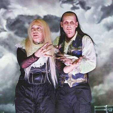 Two vampires stand close together and look seriously into the audience. They wear black trousers, linen shirts with ruffles and waistcoats with decorations. Their hair is long and unkempt, their fingers long, with long pointed fingernails. Their skin is pale, they have deep circles under their eyes. A sky with dark clouds can be seen in the background.