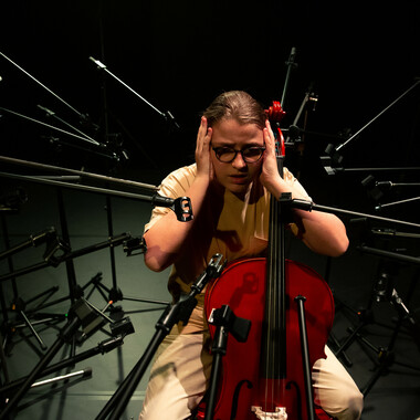 Bildbeschreibung seitens der Festival Theaterformen Redaktion. Selbstbeschreibung der abgebildeten Personen folgt. / Eine sitzende Person mit beiger Kleidung und Brille hält sich mit den Händen die Ohren zu. Zwischen ihren Beinen befindet sich ein Cello. Die Person ist in einem Kreis von leeren Mikrofonständern umgeben, die in Richtung ihres Kopfes zeigen.