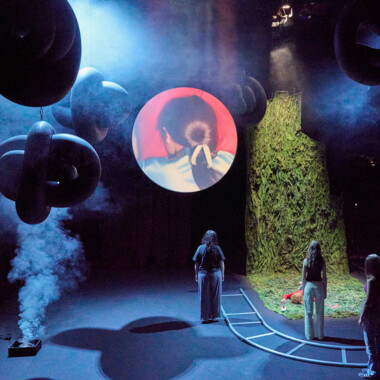 Four people are standing with their backs to us, a projection screen hangs from the ceiling, and one person is lying on the floor.
