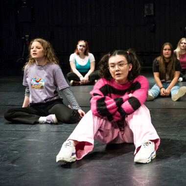 Bühnensituation: Mehrere Teenager sitzen versetzt auf dem Bühnenboden.