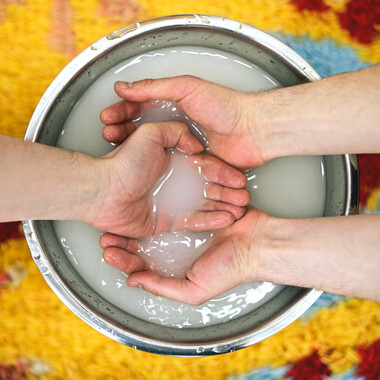 A metal bowl with a milky liquid. Three cupped hands are holding some of that liquid and slightly touching its surface.