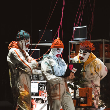 Bühnensituation: Drei maskierte Personen stehen und schauen in einen Laptop. Im Hintergrund Kabel und Regale.
