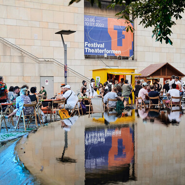 Bildbeschreibung seitens der Festival Theaterformen Redaktion. Selbstbeschreibung der abgebildeten Personen folgt. / Im Hintergrund ist die Außenfassade des Kleinen Haus mit einem Festival Theaterformen Banner zu sehen. Davor sind viele Menschen. Einige davon sitzen an den runden Tischen, einige stehen Schlange an zwei Verkaufsständen, die auf der rechten Seite sind. Der linke Stand ist gelb mit einem flachen Dach und der rechte hat einen spitzen Dach, so wie ein Häuschen und ist dunkelbraun. Im Vordergrund ist ein Brunnen zu sehen, in dem sich alles spiegelt.