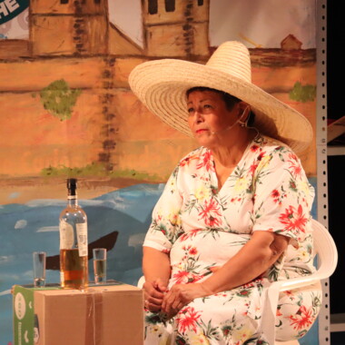 Eine ältere Frau sitzt auf einer Bühne auf einem Plastikstuhl. Sie trägt ein geblümtes Kleid und einen großen Strohhut. Neben ihr stehen eine Schnapsflasche und zwei Gläser auf einem Pappkarton, im Hintergrund ist eine gemalte Landschaft zu sehen.