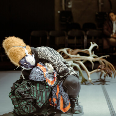 Stage situation. A masked person is crouching and clutching various fabrics. A large antler in the background.