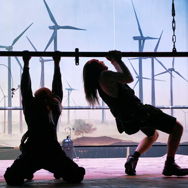Two people on a stage, clinging to a pole, they are in the shadows. To the left and right of the pole hang spotlights. In the background there is a printed curtain with a landscape and windmills.  The person on the left can be seen from behind, clinging to the pole while kneeling on the ground.  The person on the right is standing directly next to it and can be seen from the side. The person grips the pole with both hands and seems to be pulling himself up on it.