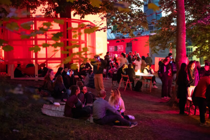 Festivalzentrum am Gartenhaus Haeckel unter freiem Himmel: Das Foto zeigt das gut besuchte Festivalzentrum bei Nacht. Einige Besucher*innen sitzen auf dem Boden, der mit rotem Hackschnitzel belegt ist, andere sitzen an Tischen oder stehen. Das Festivalzentrum ist in rotes Licht eingetaucht. 